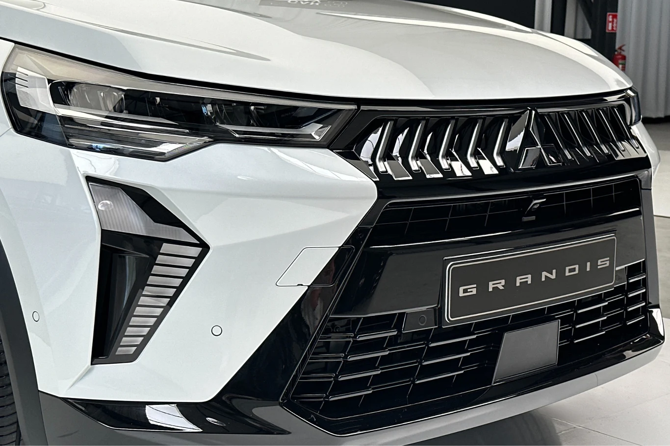 Grandis otrzymał nowe wzornictwo charakterystycznej dla Mitsubishi przedniej części nadwozia Dynamic Shield. Przód białego samochodu Grandis z nowoczesnymi reflektorami LED i masywnym czarnym grillem, logo marki umieszczone pośrodku, karoseria o wyrazistych liniach.