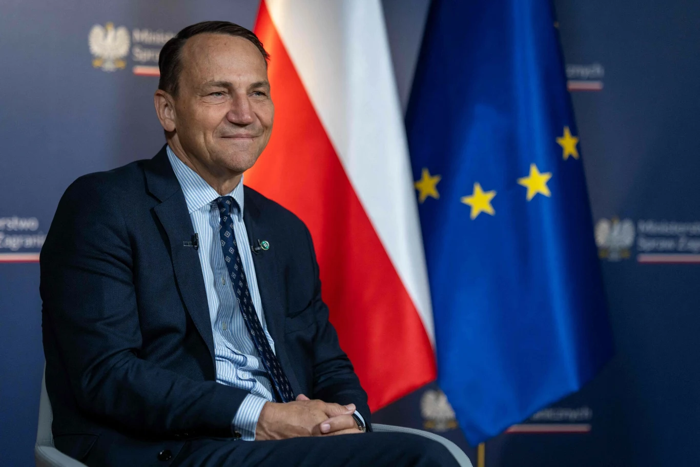 Radosław Sikorski określił tegoroczny szczyt NATO jako bardzo "owocny" Mężczyzna w garniturze siedzi na krześle na tle flag Polski oraz Unii Europejskiej, w tle widoczne oficjalne logo instytucji państwowej.