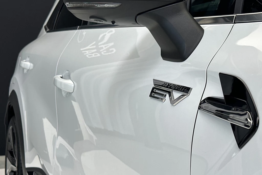 Biały nowoczesny samochód z widocznym emblematem EV świadczącym o napędzie elektrycznym, detal lusterka bocznego i fragment drzwi kierowcy, elementy karoserii lśnią w świetle
