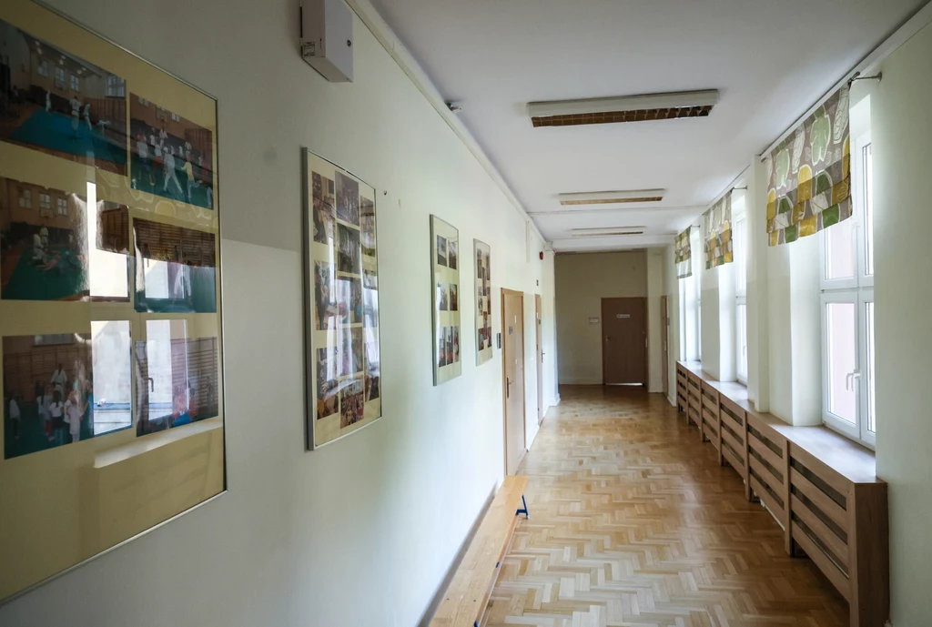 Licealiści nie kryją obaw wobec dodatkowych przedmiotów w szkole Jasny szkolny korytarz z drewnianą podłogą, dużymi oknami po jednej stronie oraz ścianą ozdobioną licznymi zdjęciami w ramkach po drugiej stronie.