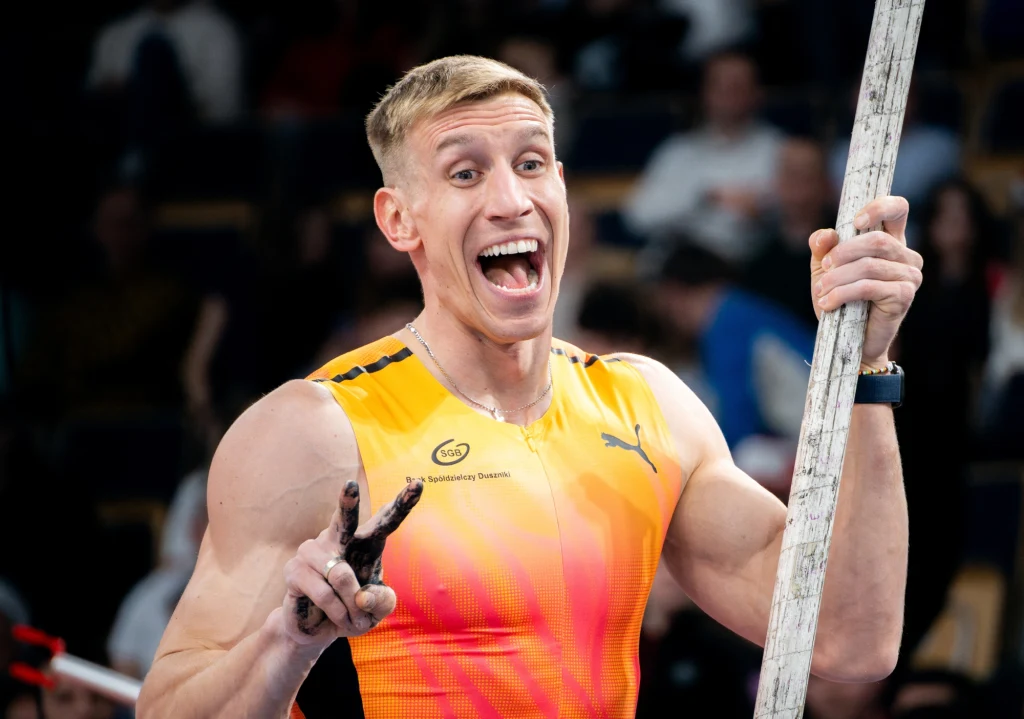 Sportowiec w pomarańczowo-żółtym stroju sportowym z wyciągniętą tyczką w ręku szeroko uśmiecha się i pokazuje znak pokoju dwoma palcami, w tle rozmyta widownia.