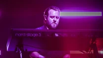 Mężczyzna grający na elektronicznych keyboardach Nord Stage 3 i Nord Stage 4, oświetlony różowym i fioletowym światłem scenicznym, skupiony na grze podczas koncertu.