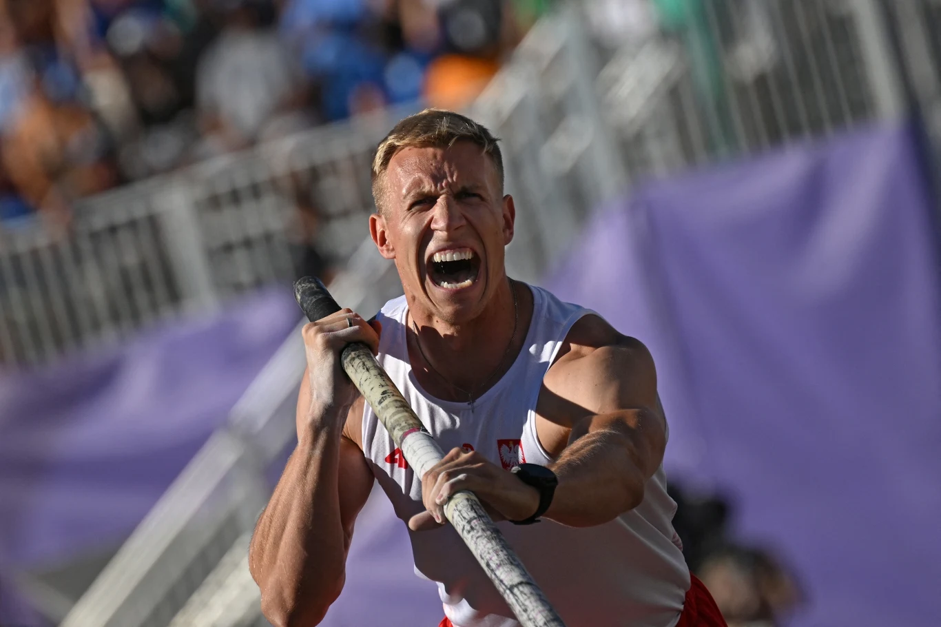 Piotr Lisek Zawodnik w stroju sportowym z intensywnym wyrazem twarzy trzymający tyczkę podczas skoku o tyczce, na trybunach w tle rozmazana publiczność