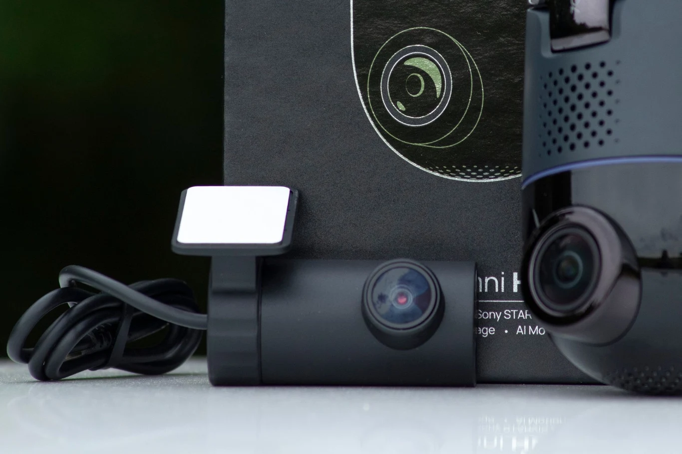 Zbliżenie na nowoczesną kamerę samochodową z widocznym obiektywem i przymocowanym kablem, obok znajduje się fragment opakowania z graficznym przedstawieniem obiektywu i inna część elektroniczna z widoczną soczewką.