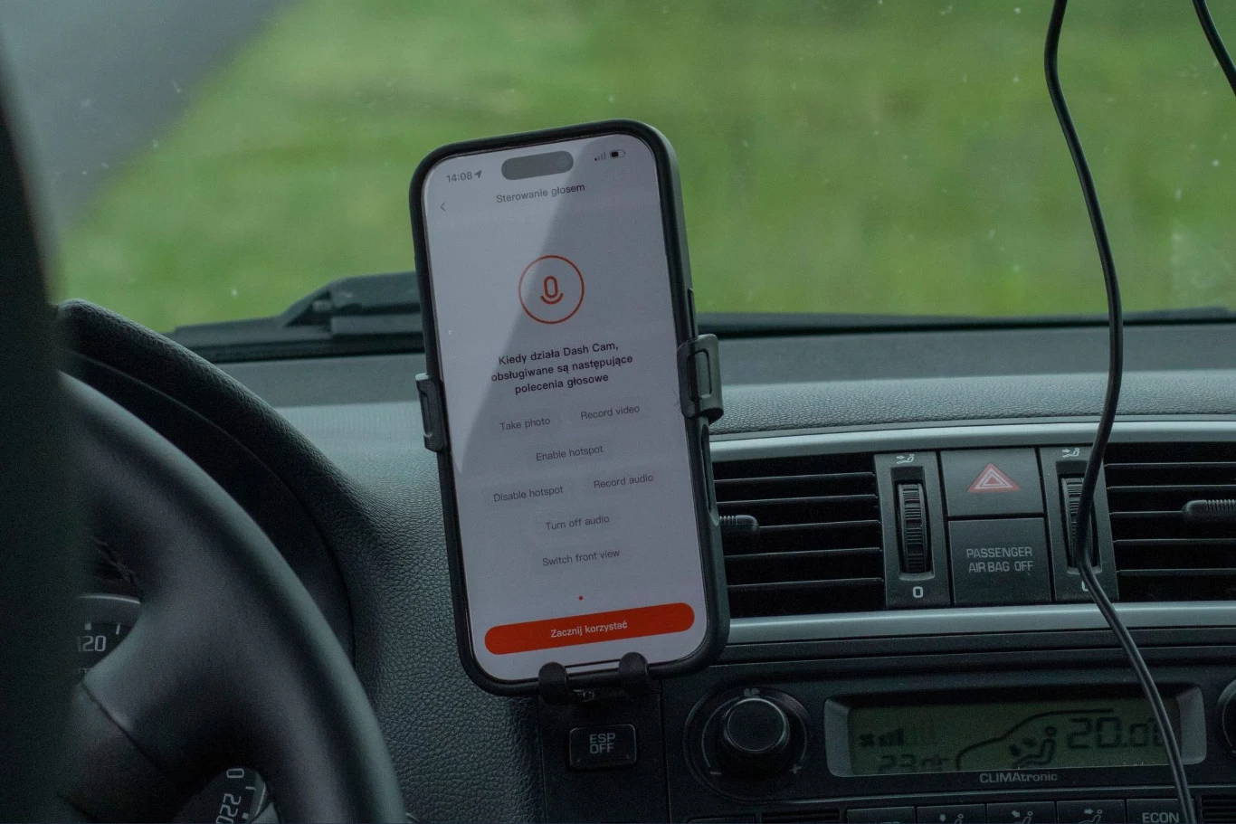 Telefon zamontowany na uchwycie w samochodzie z uruchomioną aplikacją na ekranie, widoczne fragmenty deski rozdzielczej oraz nawiewy, w tle niewyraźna szyba i zieleń.