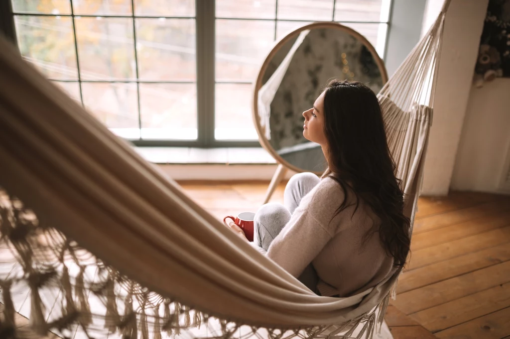 Selfcare bez presji Młoda kobieta siedzi w hamaku, wpatrując się zamyślona przez duże okno, z kubkiem w dłoni, w tle duże lustro opierające się o ścianę i drewniana podłoga