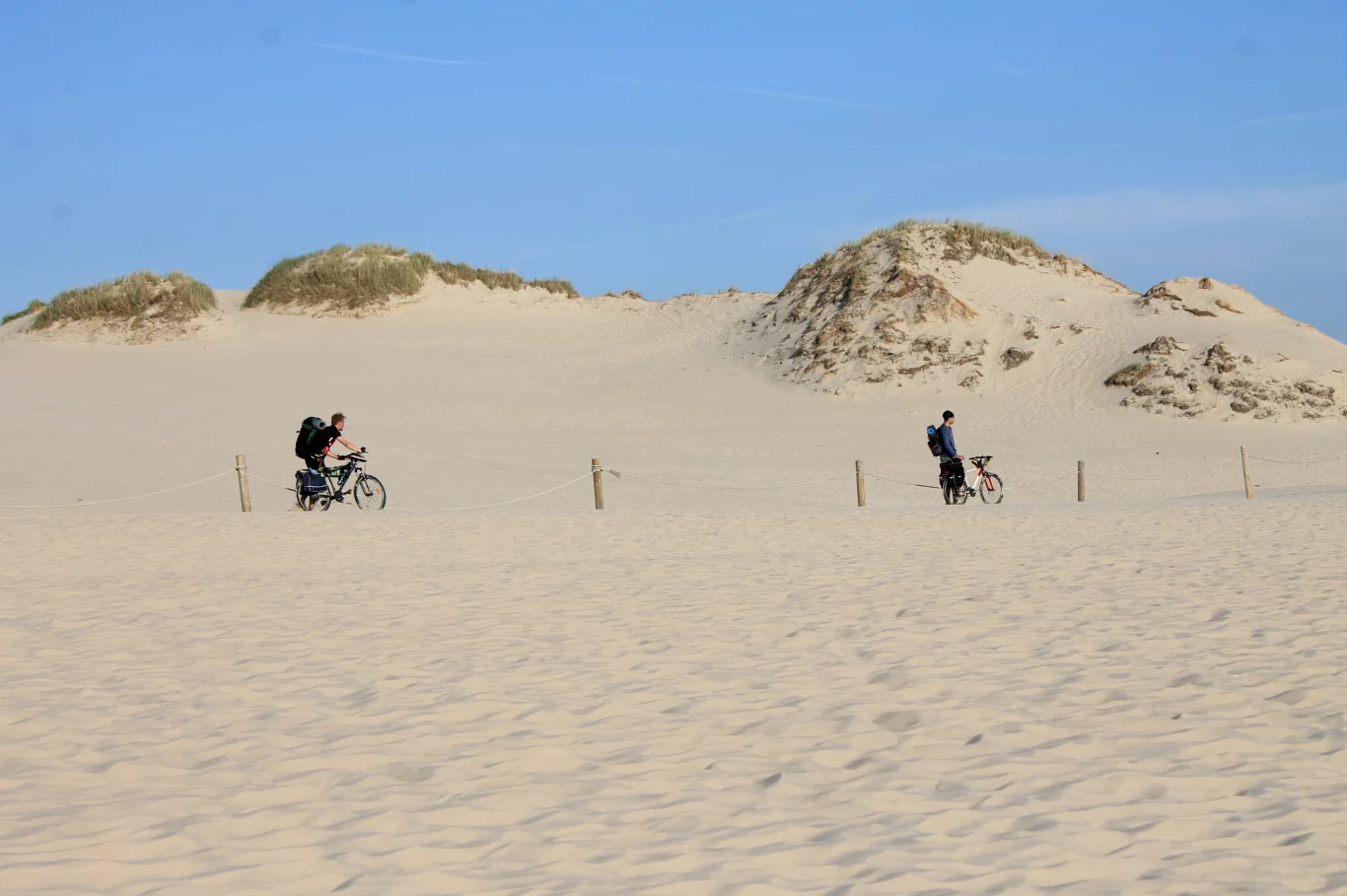 Dwóch rowerzystów przemierza piaszczystą plażę w otoczeniu jasnych wydm i błękitnego nieba, tworząc spokojną, letnią atmosferę.