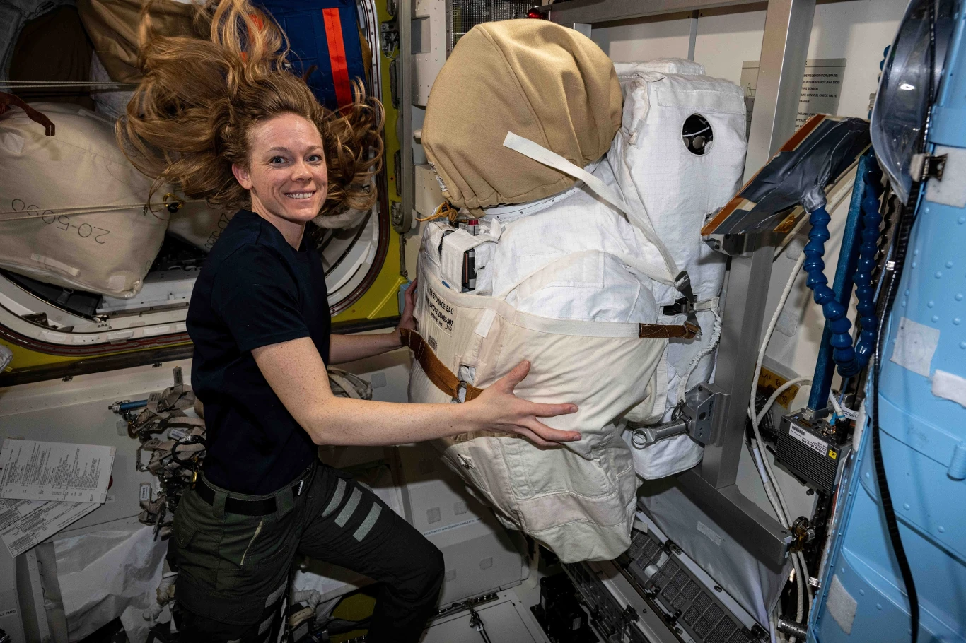 Astronautka z unoszącymi się włosami w stanie nieważkości trzyma biały skafander kosmiczny wewnątrz modułu stacji kosmicznej, otoczona sprzętem i panelami technicznymi.