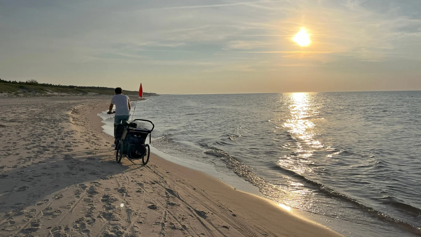 Mężczyzna prowadzący rower z przyczepką rowerową wzdłuż spokojnej, piaszczystej plaży podczas zachodzącego słońca, fale delikatnie obmywają brzeg, a na horyzoncie widoczna jest linia lasu.