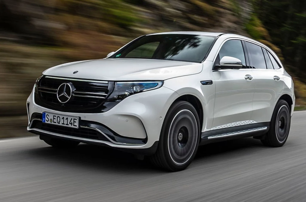 Artur Łącki (KO) ma elektrycznego Mercedesa EQC. Biały samochód typu SUV marki Mercedes-Benz EQC jedzie po asfaltowej drodze, pośród zielonej roślinności i skał w tle, w ruchu widoczna dynamiczna jazda po zakręcie.