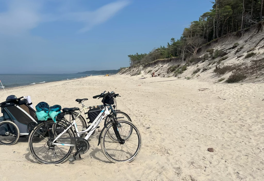 Wybierając się w rowerową podróż przez Słowiński Park, można delektować się pustymi plażami o białym miękkim piasku Dwa rowery stoją na piaszczystej plaży przy lesie, obok znajduje się przyczepka rowerowa, w tle spokojne morze i zalesione wydmy.