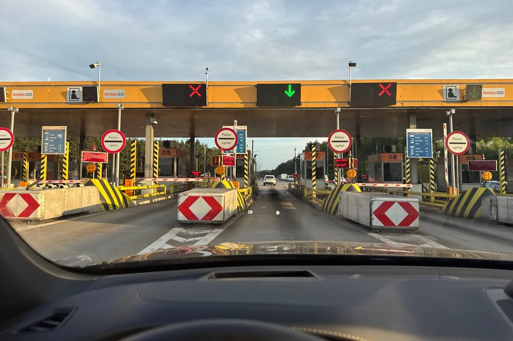 W te wakacje bramki nie będą otwarte cały czas Bramki poboru opłat na autostradzie z widocznymi znakami nakazów i zakazów, kolorowymi światłami nad przejazdami oraz pojazdami oczekującymi na przejazd; zdjęcie wykonane z wnętrza samochodu.