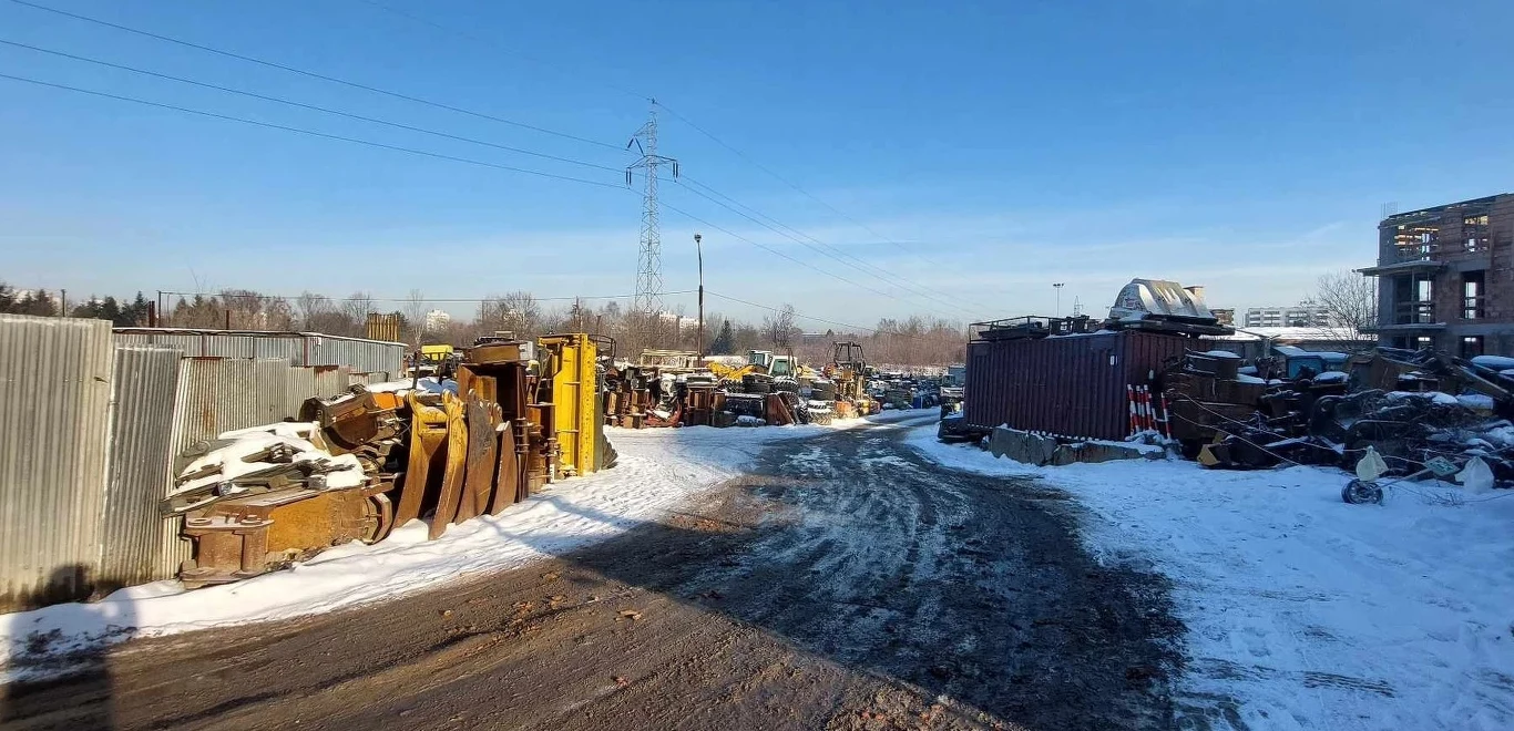 Złomowisko maszynowe z licznymi częściami i elementami dużych pojazdów oraz sprzętu ustawionymi wzdłuż ogrodzenia, ośnieżona droga, blaszany kontener i rozproszone budynki w tle pod błękitnym niebem.