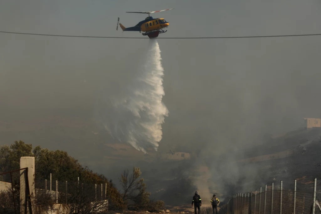Helikopter zrzuca wodę nad płonącym terenem, dwoje strażaków porusza się w stronę pożaru, wokół unosi się dym.