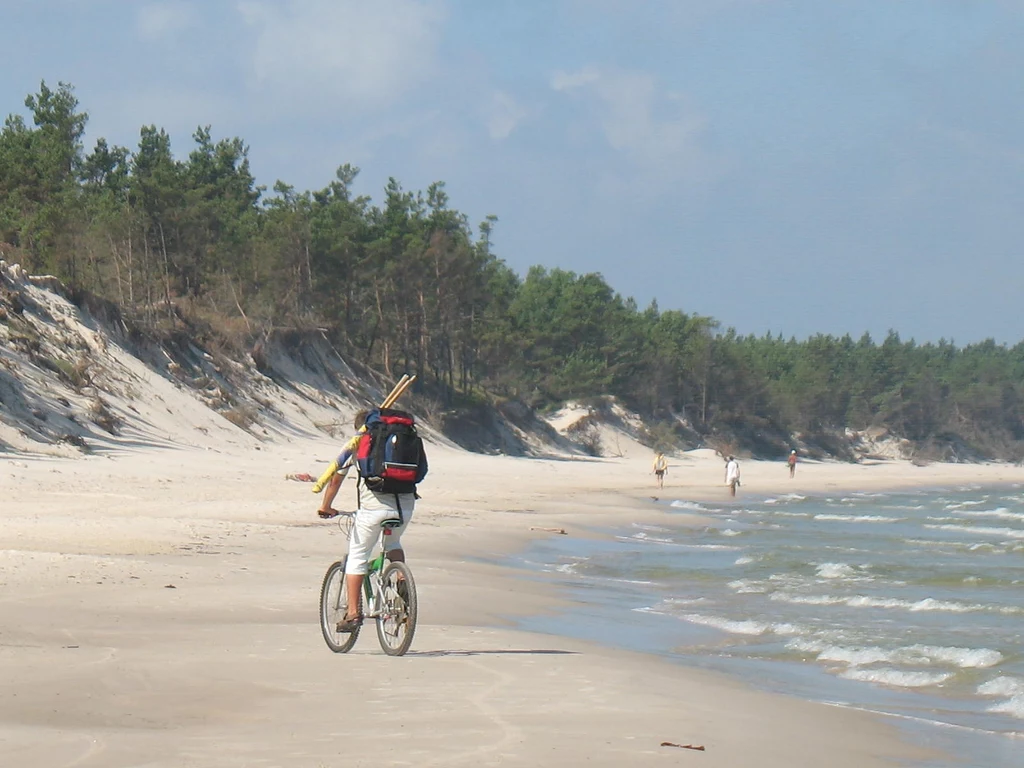 Podróż rowerem przez plaże Słowińskiego Parku Narodowego to nieoczywista bałtycka przygoda osoba z plecakiem jedzie rowerem brzegiem morza, na tle piaszczystej plaży z lasem sosnowym, w oddali widoczni są spacerowicze