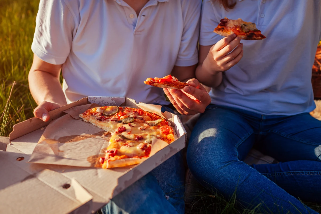Jakie jedzenie szkodzi latem Dwie osoby siedzą na trawie i jedzą pizzę z kartonowego pudełka, obie ubrane w białe koszulki i niebieskie dżinsy, ciepłe popołudniowe światło podkreśla atmosferę swobodnego pikniku.