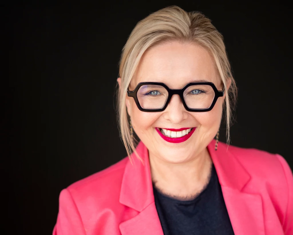 Iwona Wencel, HR Executive z ponad 25-letnim doświadczeniem Uśmiechnięta kobieta o blond włosach, nosząca duże czarne okulary i różowy żakiet na ciemnym tle.