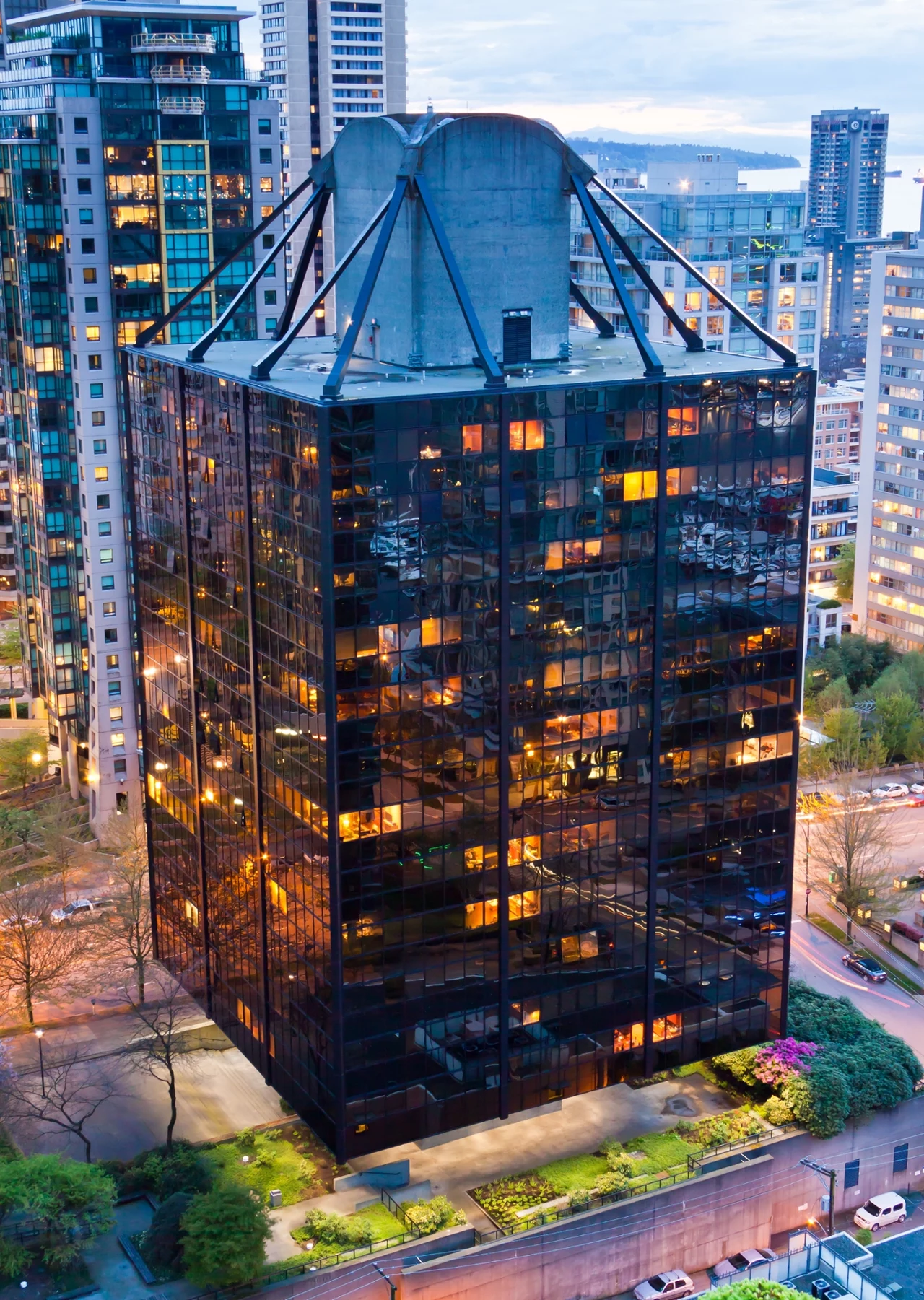 Zdjęcie budynku The Qube w Vancouver w Kanadzie. Czy tak mógłby wyglądać Trzonolinowiec? Nowoczesny biurowiec o przeszklonej elewacji otoczony zielenią i znajdujący się wśród wysokich budynków w centrum miasta, wnętrza rozświetlone światłem, a na dachu widoczna nietypowa konstrukcja.