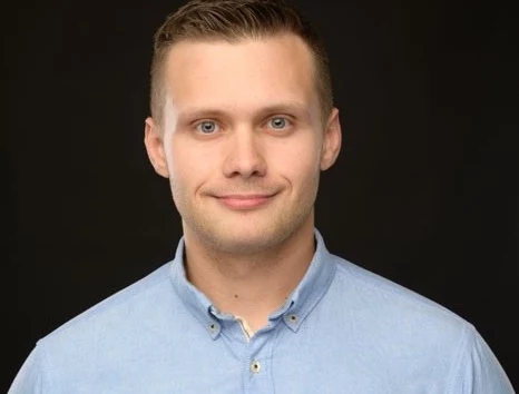 Franciszek Ostaszewski, Psycholog, słuchacz Interdyscyplinarnej Szkoły Doktorskiej Uniwersytetu SWPS Młody mężczyzna w jasnoniebieskiej koszuli patrzy prosto w obiektyw, delikatnie się uśmiecha, ciemne tło podkreśla sylwetkę i wyraz twarzy.