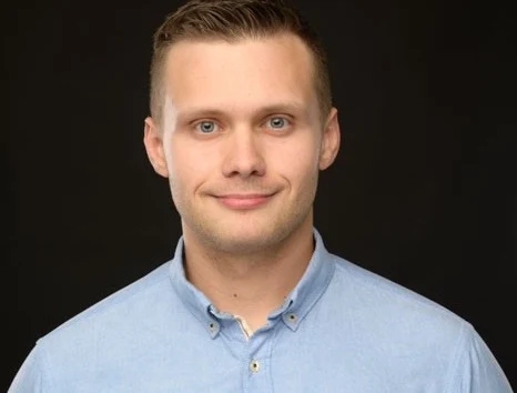 Młody mężczyzna w jasnoniebieskiej koszuli patrzy prosto w obiektyw, delikatnie się uśmiecha, ciemne tło podkreśla sylwetkę i wyraz twarzy.