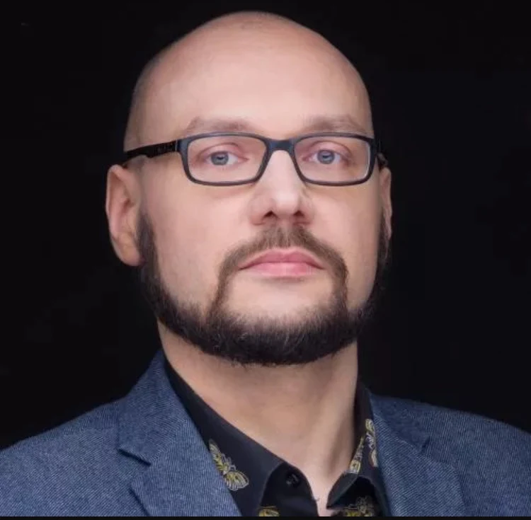 Łysy mężczyzna z brodą i okularami, ubrany w ciemną marynarkę i koszulę, patrzący prosto w obiektyw na ciemnym tle.