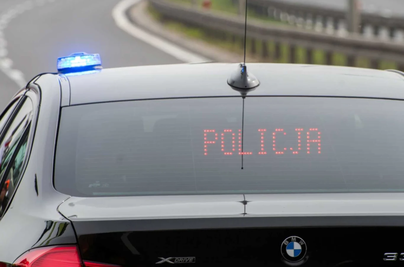 Radiowóz policyjny marki BMW z włączonym niebieskim sygnałem świetlnym oraz elektronicznym wyświetlaczem z napisem 'POLICJA' jadący po asfaltowej drodze dwujezdniowej.