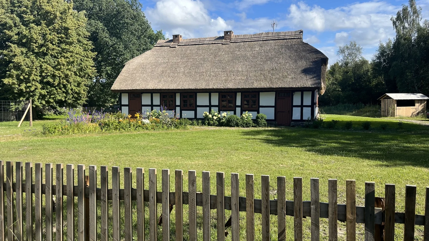 Drewniany dom kryty strzechą otoczony zielonym trawnikiem i drewnianym płotem, po prawej stronie widoczna szopa, w tle wysokie drzewa i niebieskie niebo z białymi chmurami.