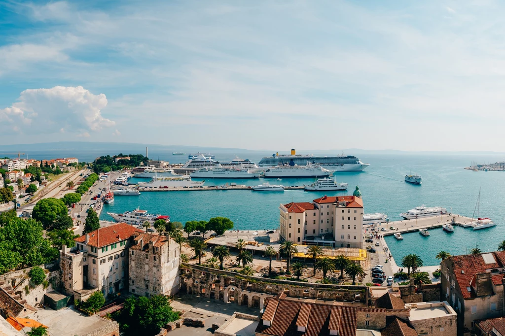 Kierunki na urlop poza sezonem Widok na port z cumującymi dużymi statkami wycieczkowymi, kilka mniejszych łodzi, zabudowa miejska w pierwszym planie oraz malownicze wybrzeże i spokojne, błękitne morze w tle.
