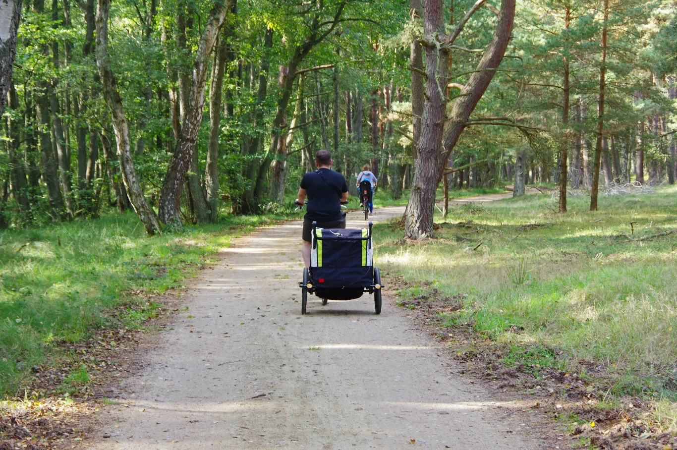 Leśna ścieżka prowadzi przez gęsty las, po której jedzie rowerzysta z przyczepką, a w oddali widoczny drugi rowerzysta, otoczenie wypełniają letnie promienie słońca i zielone drzewa.