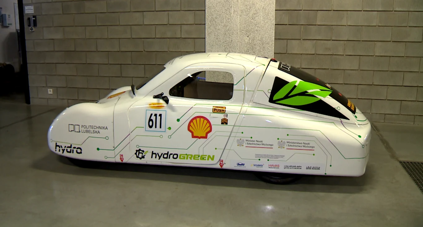 Białe, aerodynamiczne auto testowe z napędem wodorowym stojące w garażu, na karoserii widoczne logotypy sponsorów i uczelni technicznej oraz elementy graficzne nawiązujące do ekologii i nowych technologii.