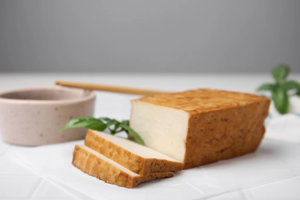 Nie smakuje ci naturalne tofu? Spróbuj wędzone, które przypomina trochę serki góralskie typu oscypek. Jest gotowe do użycia, wystarczy pokroić na plasterki i położyć na kanapkę jak plastry sera Kostka wędzonego tofu pokrojona na plastry, obok niewielka miseczka i świeże listki bazylii na białym tle.