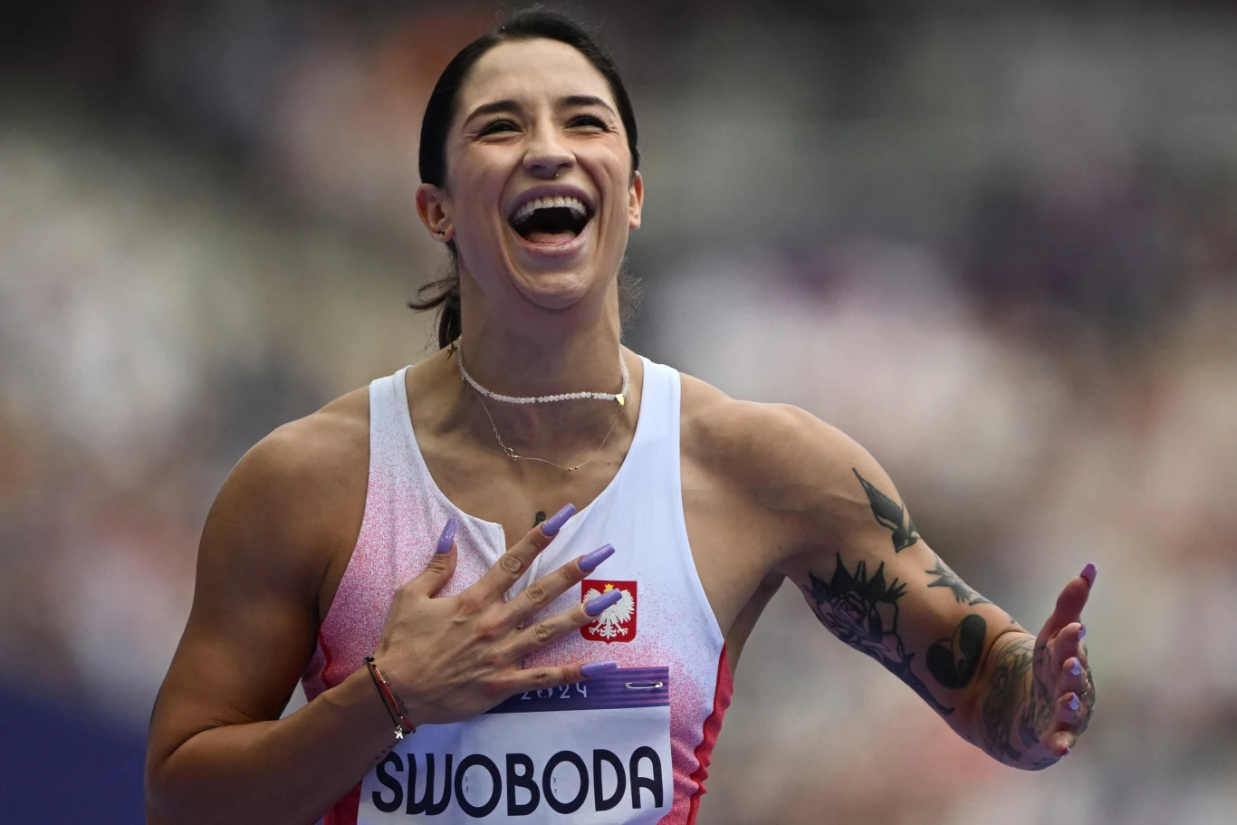 Zawodniczka lekkoatletyczna w sportowym stroju z numerem startowym i nazwiskiem Swoboda na piersi, szeroko uśmiechnięta, z ręką położoną na klatce piersiowej, wyrażająca radość i emocje po starcie na stadionie.