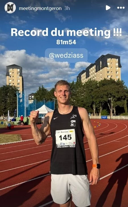 Uśmiechnięty sportowiec w sportowej koszulce z numerem startowym 145 stoi na bieżni lekkoatletycznej, unosząc zaciśniętą pięść w geście triumfu, w tle widoczne są bloki mieszkalne, zieleń i elementy stadionu.
