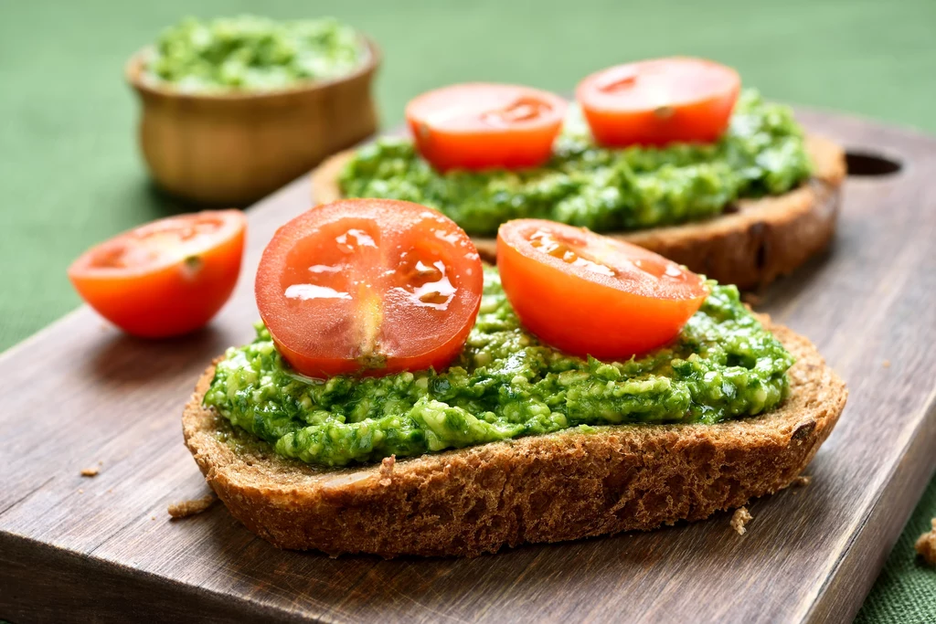 Pasta z bobu świetnie smakuje na grzankach Kromki chleba posmarowane zielonym pesto, udekorowane połówkami świeżych pomidorów, ułożone na drewnianej desce, w tle miseczka z dodatkową porcją zielonej pasty.