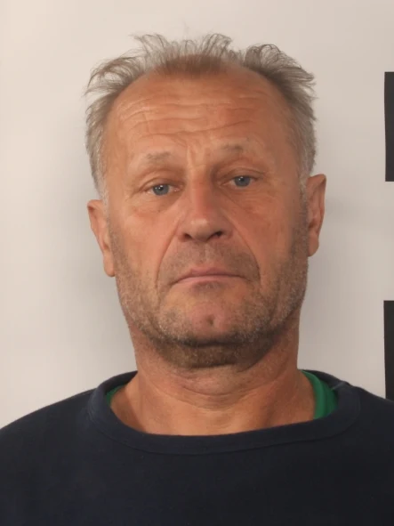 Mężczyzna w średnim wieku z krótkimi, przerzedzonymi włosami i kilkudniowym zarostem spogląda prosto w obiektyw na jasnym tle