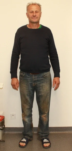 Mężczyzna w średnim wieku stoi na tle jasnej ściany, ubrany w ciemny sweter, znoszone jeansy z widocznymi zabrudzeniami oraz klapki, z delikatnym uśmiechem na twarzy.