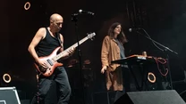 Dwóch muzyków na scenie koncertowej, mężczyzna gra na gitarze basowej, kobieta obsługuje klawisze, ciemne tło z elementami oświetlenia scenicznego, widoczne mikrofony i sprzęt muzyczny.