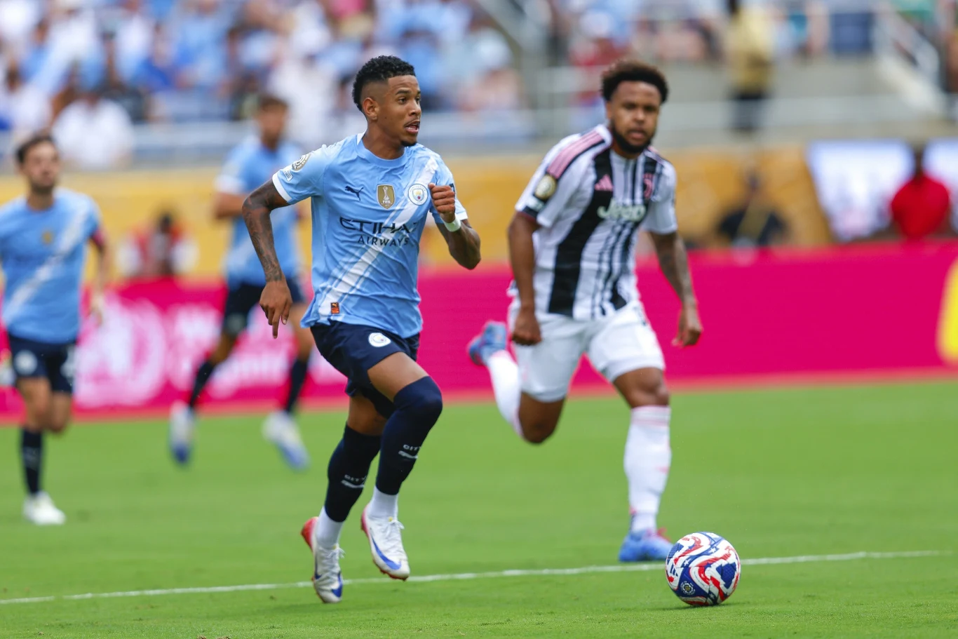Dwóch piłkarzy biegnących z piłką po zielonym boisku, jeden ubrany w niebieski strój z logo klubu Manchester City, ścigany przez zawodnika w biało-czarnej koszulce, w tle rozmazana publiczność na trybunach.