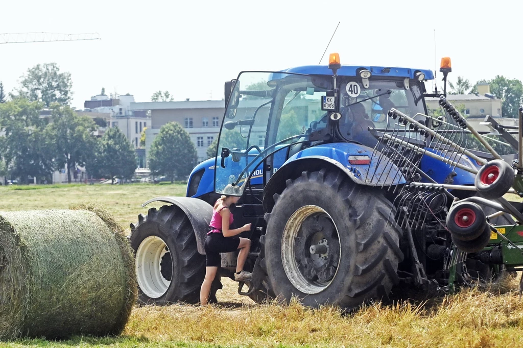 Duży traktor rolniczy na polu podczas pracy, blisko stoi kobieta w krótkich spodenkach wsiadająca do pojazdu, z przodu widoczna belka siana, w tle zabudowania miejskie i drzewa.