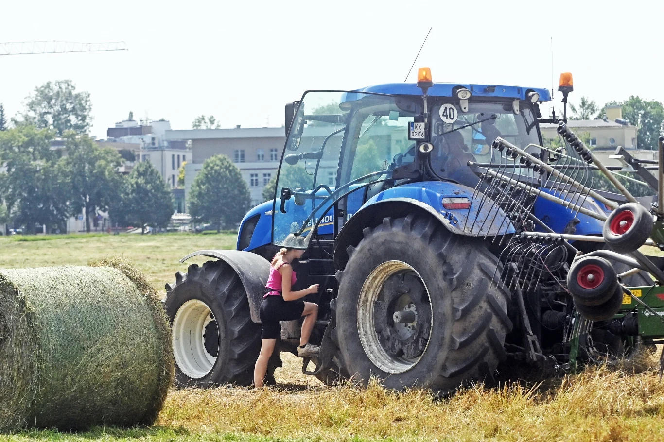 Duży traktor rolniczy na polu podczas pracy, blisko stoi kobieta w krótkich spodenkach wsiadająca do pojazdu, z przodu widoczna belka siana, w tle zabudowania miejskie i drzewa.