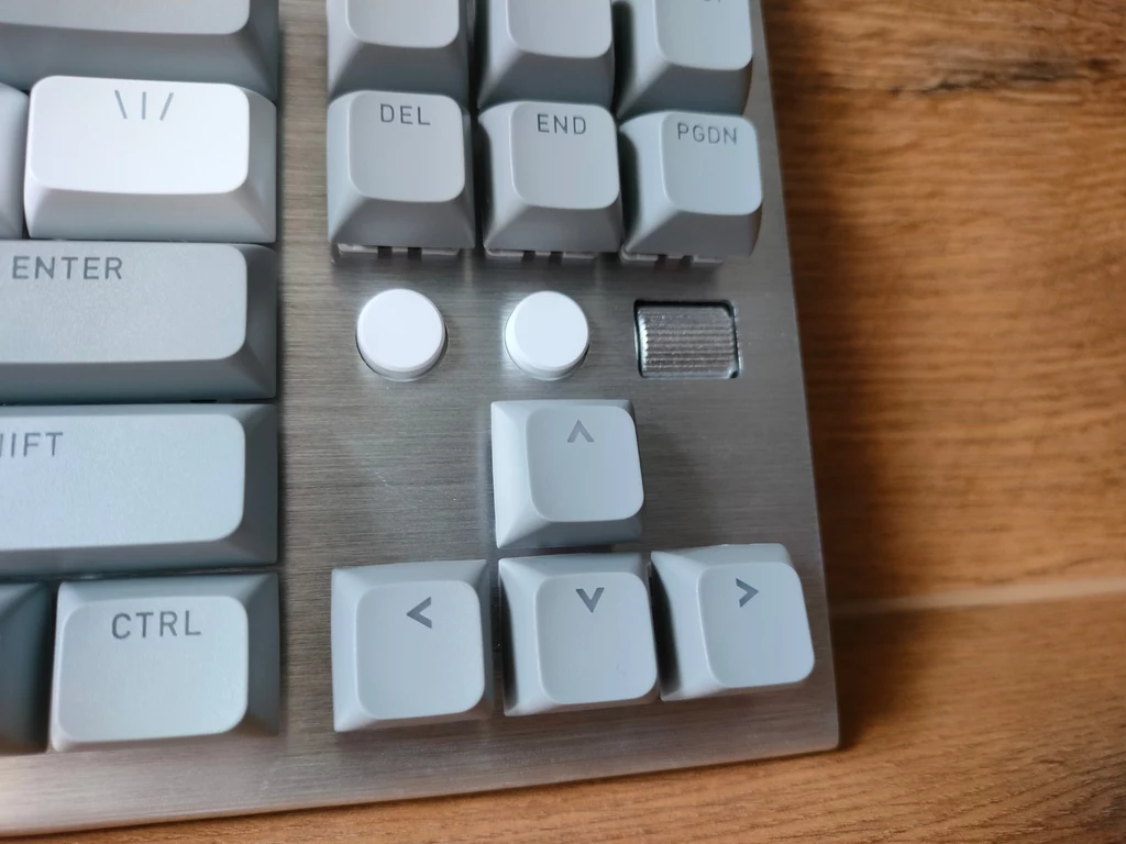 Hator Gravity TKL Fragment klawiatury komputerowej z widocznymi klawiszami strzałek, klawiszami funkcyjnymi DEL, END i PGDN oraz dwoma okrągłymi przyciskami i rolką na metalowej powierzchni obudowy. Tło stanowi drewniany blat.