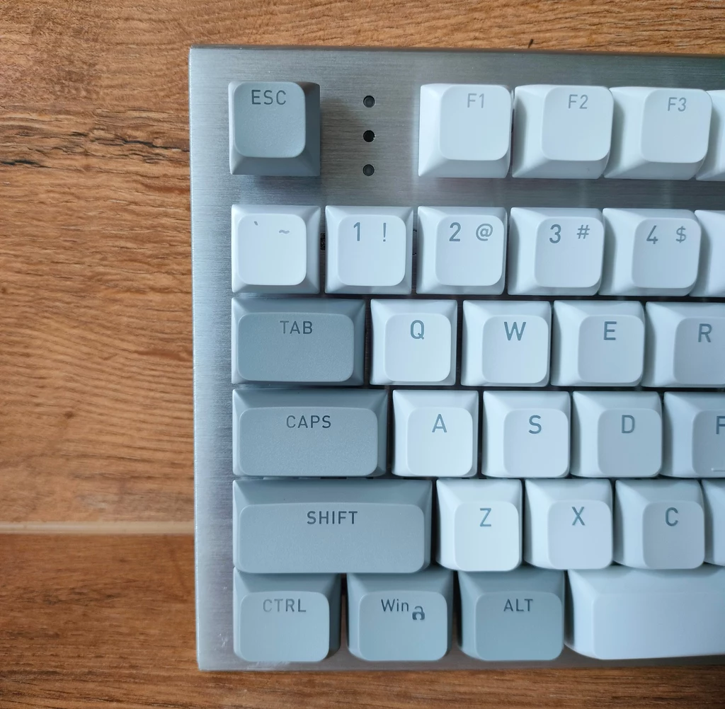 Hator Gravity TKL Fragment klawiatury mechanicznej z białymi klawiszami na tle drewnianej powierzchni, widoczne są klawisze funkcyjne oraz litery i symbole w lewej górnej części klawiatury.