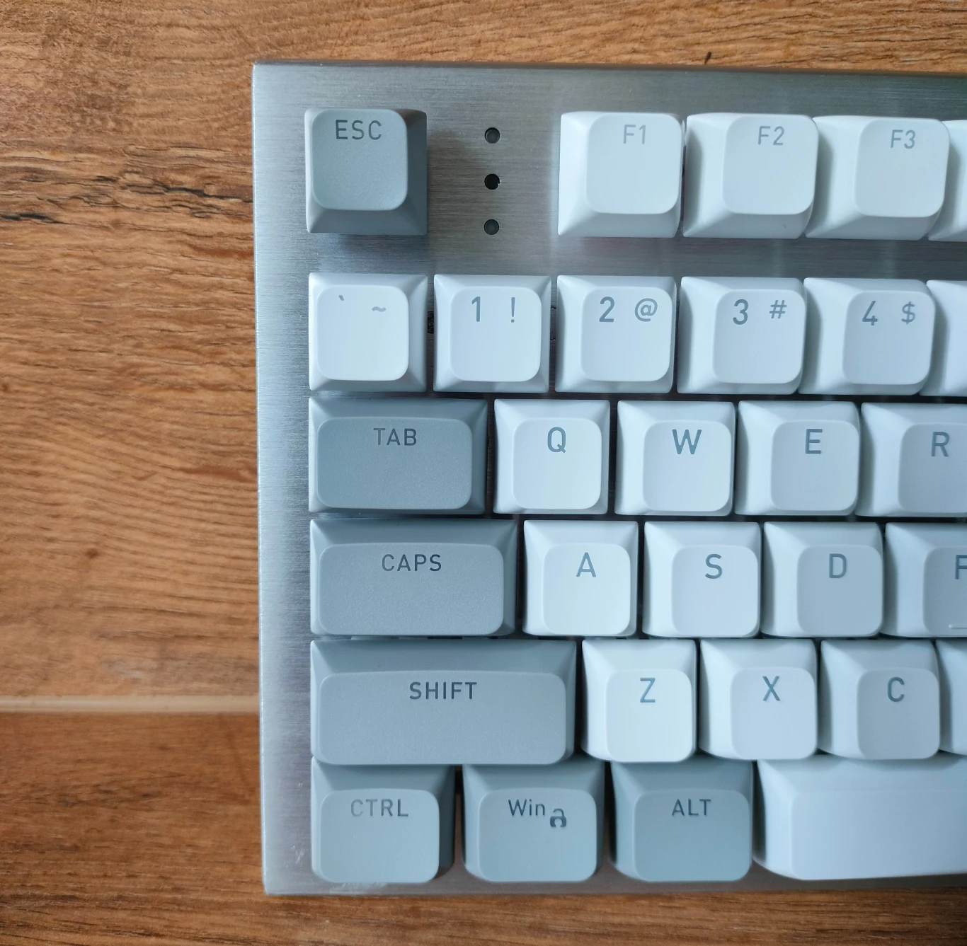 Fragment klawiatury mechanicznej z białymi klawiszami na tle drewnianej powierzchni, widoczne są klawisze funkcyjne oraz litery i symbole w lewej górnej części klawiatury.