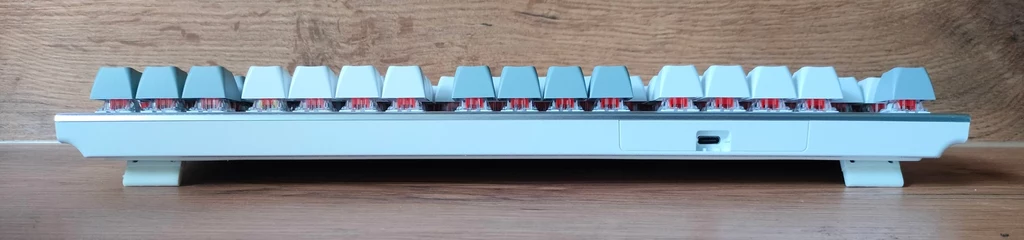 Hator Gravity TKL Mechaniczna klawiatura komputerowa widziana z boku, z widocznymi podświetlanymi przełącznikami i klawiszami w odcieniach szarości, ustawiona na drewnianej powierzchni na tle drewnianej ściany.