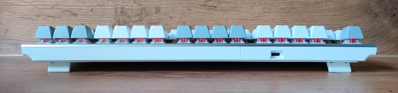 Mechaniczna klawiatura komputerowa widziana z boku, z widocznymi podświetlanymi przełącznikami i klawiszami w odcieniach szarości, ustawiona na drewnianej powierzchni na tle drewnianej ściany.