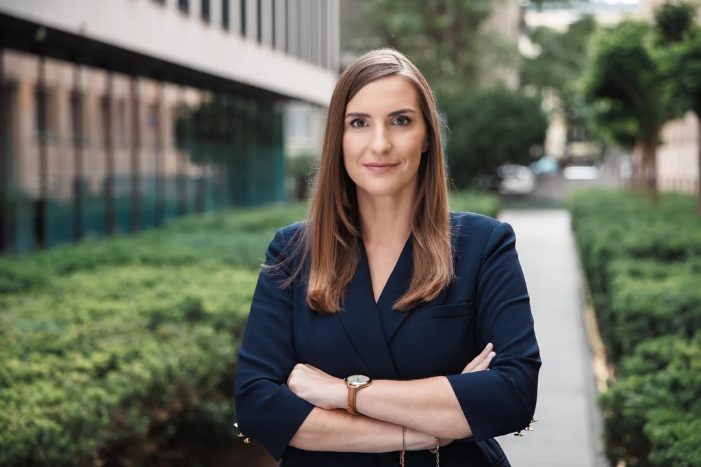 Magdalena Sobkowiak-Czarnecka, wiceminister ds. Unii Europejskiej Kobieta w średnim wieku w granatowej marynarce z długimi, prostymi włosami stoi z założonymi rękami na tle nowoczesnych budynków i zieleni miejskiej alei.