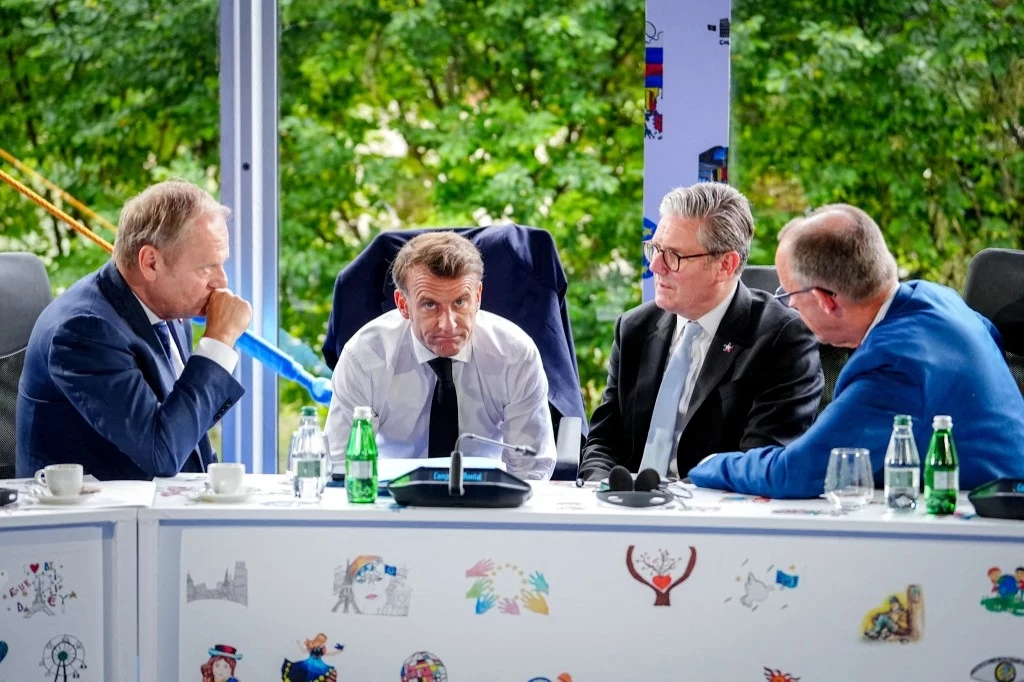 Od lewej: Donald Tusk (premier Polski), Emmanuel Macron (prezydent Francji), Keir Starmer (premier Wielkiej Brytanii), Friedrich Merz (kanclerz Niemiec) Czterech mężczyzn w garniturach siedzi przy stole dyskutując, przed nimi stoją butelki wody, szklanki oraz dokumenty. W tle widoczne zielone drzewa za przeszkloną ścianą.