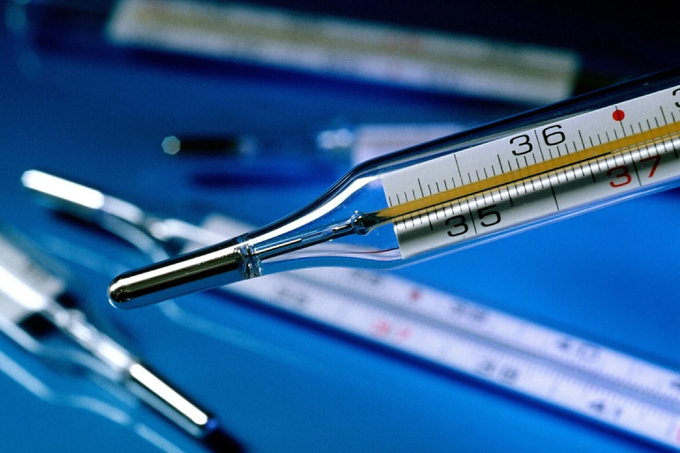 Szklany termometr rtęciowy z czytelną skalą temperatury na niebieskim tle, w tle widoczne rozmyte inne termometry.