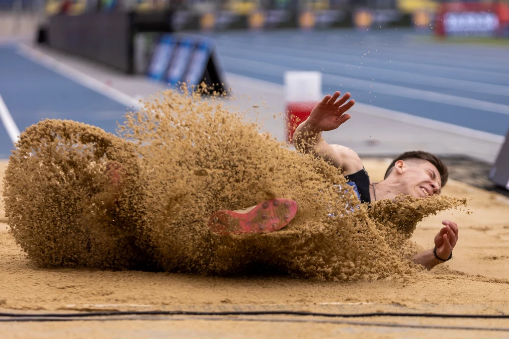 Sportowiec w trakcie lądowania podczas skoku w dal, piasek unosi się intensywnie wokół jego ciała, widoczne są dynamiczne ruchy i wysiłek fizyczny zawodnika.