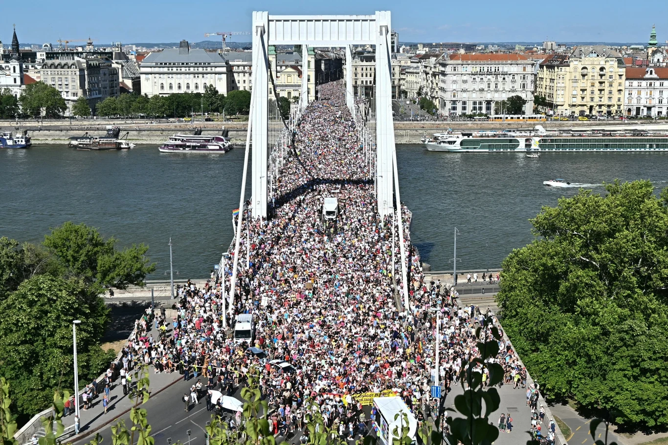 Parada równości w Budapeszcie. Wcześniej zakazała jej policja Tłum ludzi maszerujący przez most na tle szerokiej rzeki, w otoczeniu miejskich budynków i zieleni, na niebie bezchmurna pogoda, panorama miasta widoczna w tle.